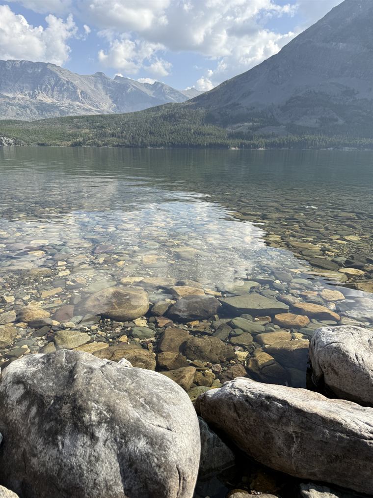 Rounded coloured stones on the lake bed, clearly visible through shallow water, with mountains rising behind.