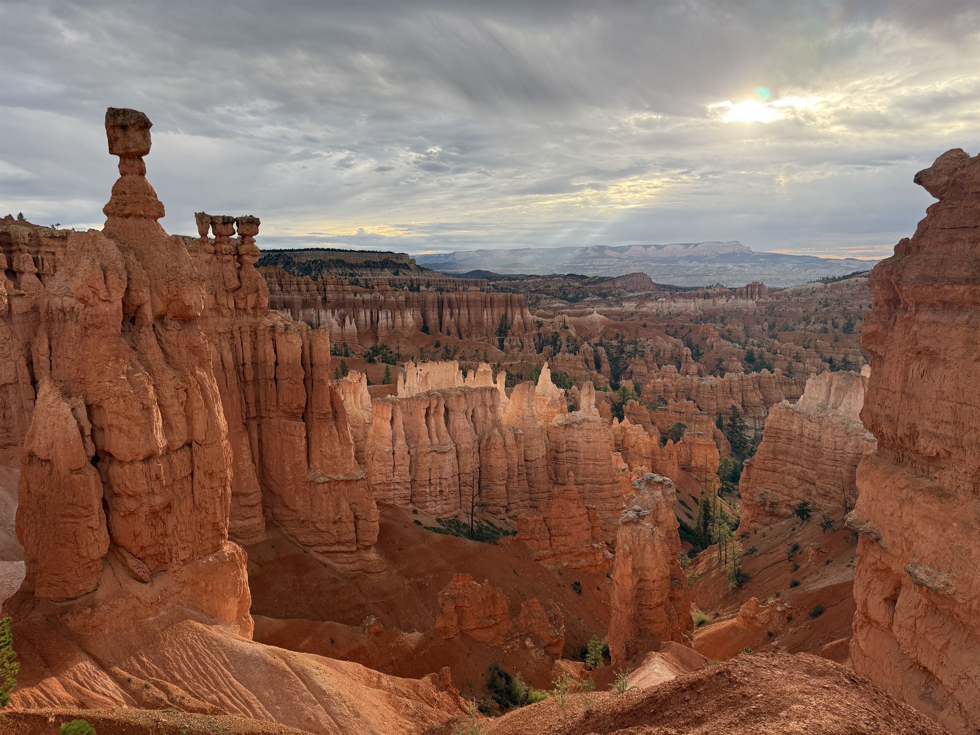 Sunlight breaking through heavy cloud over the hoodoo amphitheater at Bryce Canyon, a tall balanced-rock spire standing in the foreground.